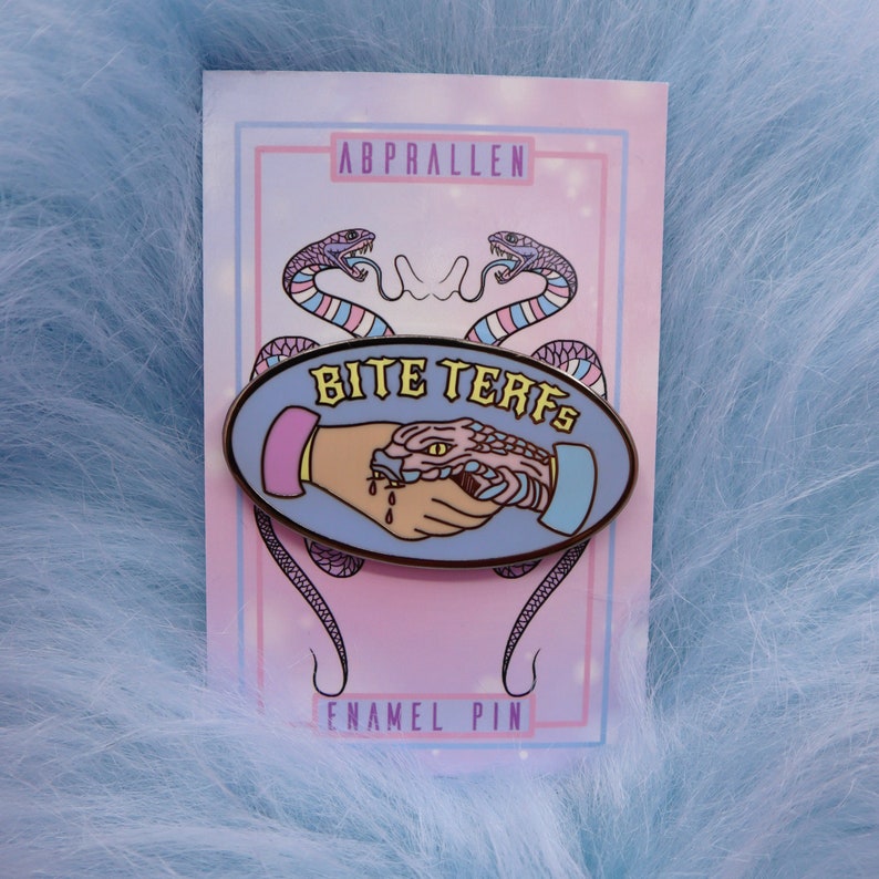 Puede incluir: Un pin de esmalte azul con una serpiente mordiendo una mano. El pin tiene forma ovalada y tiene el texto "Bite Terfs" en letras amarillas. El pin est&aacute; en una tarjeta rosa y azul con el texto "Abprallen" y "Enamel Pin".