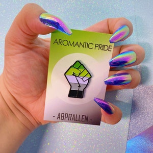 May include: An aromantic pride pin featuring a green, white, grey and black fist raised in the air. The pin is displayed on a white card with the text "Aromantic Pride" and "- ABPRALLEN -" in black.