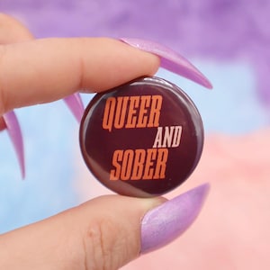 May include: A brown button with the text "QUEER AND SOBER" in orange letters.