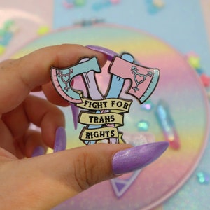 May include: A pink and blue enamel pin with two crossed axes and the text "Fight for Trans Rights".