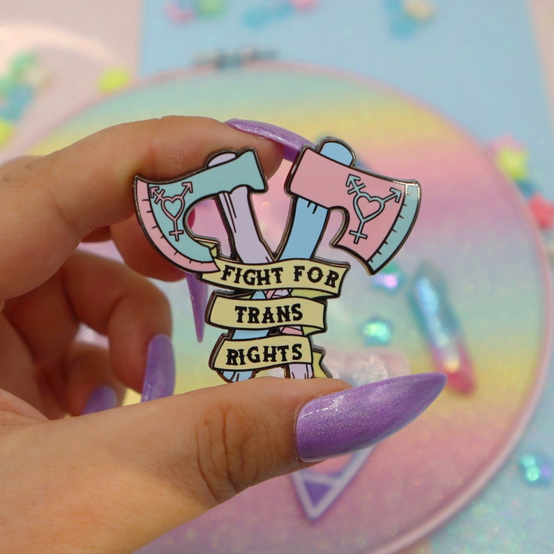 May include: A pink and blue enamel pin with two crossed axes and the text "Fight for Trans Rights".
