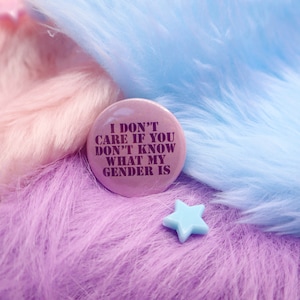 May include: A pink button with the text "I don't care if you don't know what my gender is" in purple letters. The button is on a pink and blue fuzzy surface.