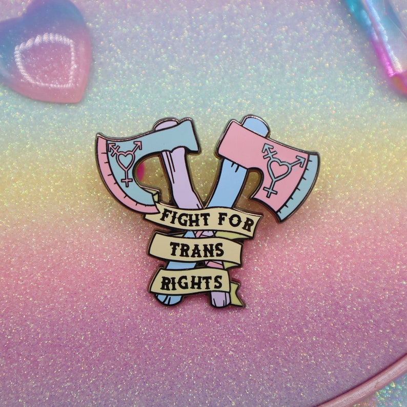 May include: A pink and blue enamel pin with two crossed axes. The axes have a transgender symbol on them. The pin has a yellow banner that says "Fight for Trans Rights".