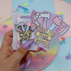 May include: Two enamel pins featuring crossed axes with a heart and the text "Fight for Trans Rights". The pins are on a card with the same text and a pink and blue color scheme.