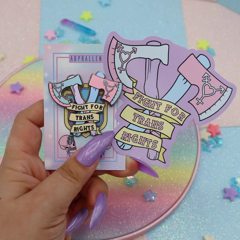 May include: Two enamel pins featuring crossed axes with a heart and the text "Fight for Trans Rights". The pins are on a card with the same text and a pink and blue color scheme.