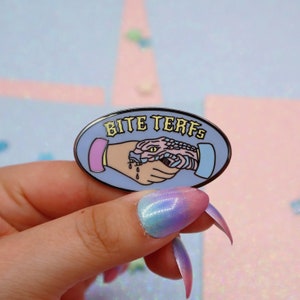 Puede incluir: Un pin de esmalte azul con una serpiente y una mano d&aacute;ndose la mano. El texto en el pin dice "Bite Terfs".