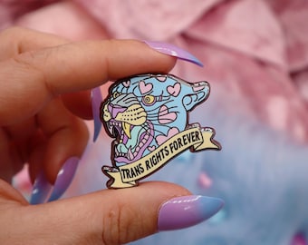 May include: A blue and pink enamel pin featuring a cartoon panther with hearts and the text "Trans Rights Forever" on a yellow banner.