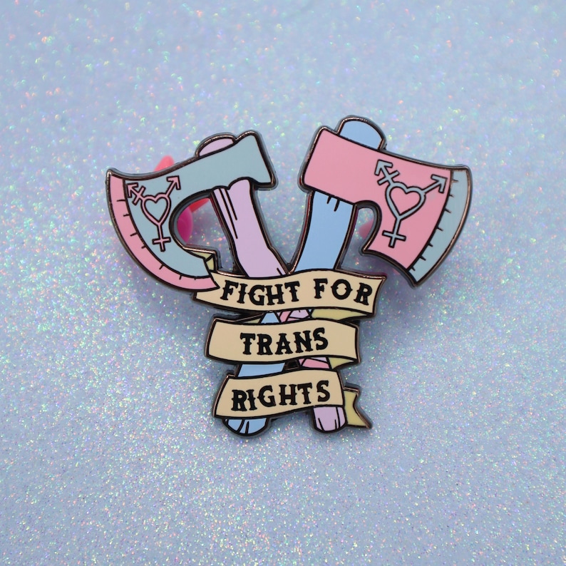 May include: A pink and blue enamel pin with two crossed axes and the text "Fight for Trans Rights".