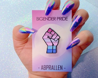 May include: A pink, blue, and white enamel pin with a raised fist design. The pin says "Bigender Pride" and "Abprallen" on a pink and blue background.