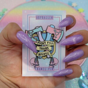 May include: A pink and blue enamel pin with two crossed axes, a transgender symbol on each axe, and a banner that reads "Fight for Trans Rights". The pin is on a white card with the words "Abprallen Enamel Pin".