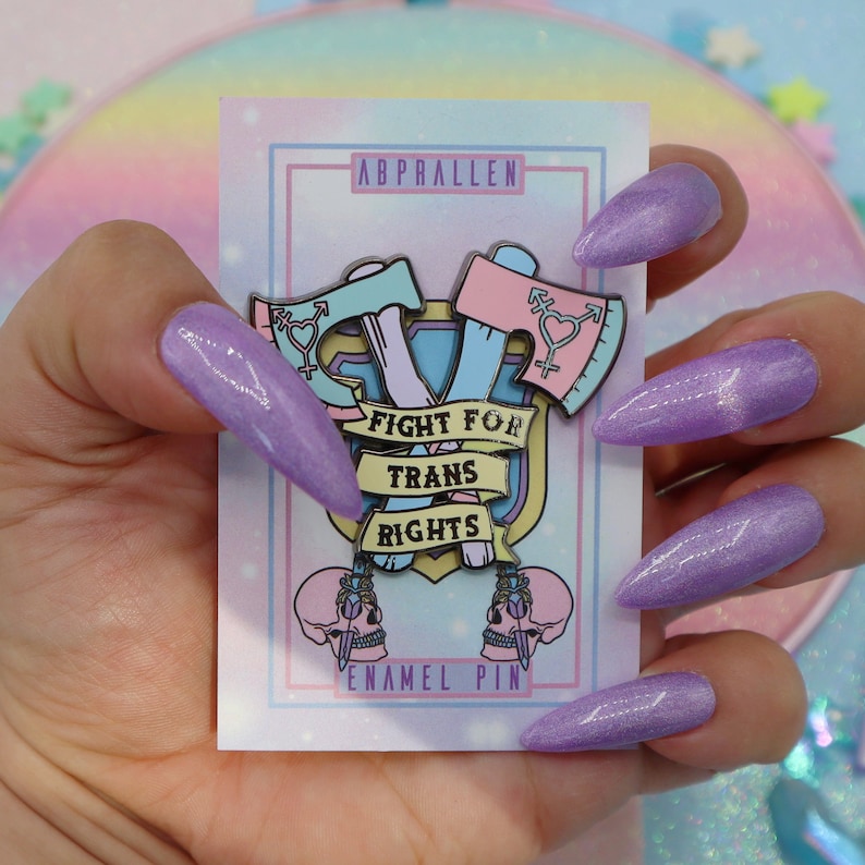 May include: A pink and blue enamel pin with two crossed axes, a transgender symbol on each axe, and a banner that reads "Fight for Trans Rights". The pin is on a white card with the words "Abprallen Enamel Pin".