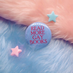 May include: A light blue badge with the red text "READ MORE GAY BOOKS". Two star-shaped decorations, one pink and one light blue, are also visible. The badge and stars are placed on a fluffy, two-tone pink and blue surface.