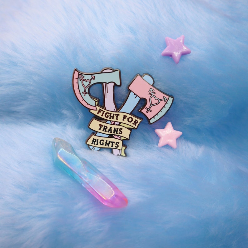 May include: A pink and blue enamel pin with two crossed axes and a banner that reads "Fight for Trans Rights". The pin is on a light blue fuzzy surface with two pink star-shaped embellishments.
