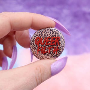 May include: A round button with a leopard print background and the words "QUEER FILTH" in red lettering.