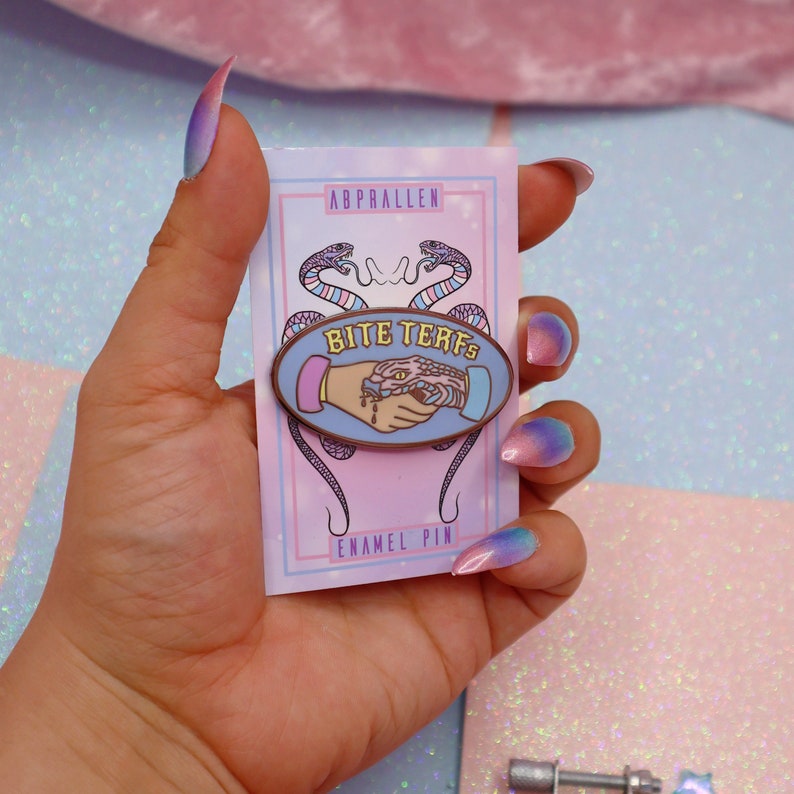 Puede incluir: Un pin de esmalte rosa y azul con una serpiente mordiendo una mano. El pin dice "Bite Terfs" en una fuente rosa y amarilla. El pin est&aacute; en una tarjeta que dice "Abprallen Enamel Pin".