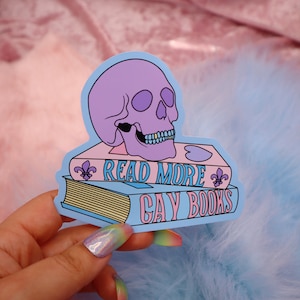May include: A sticker featuring a purple skull with a gold grill, perched atop two books. The top book is pink and reads "Read More". The bottom book is blue and reads "Gay Books".