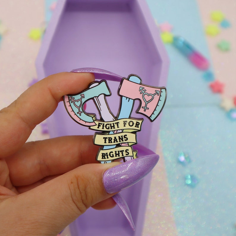 May include: A pink and blue enamel pin with two crossed axes, a transgender symbol on each axe, and a banner that reads "Fight for Trans Rights".