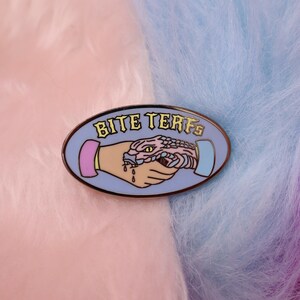 Puede incluir: Un pin de esmalte azul y rosa con una serpiente mordiendo una mano. El pin dice "Bite Terfs" en letras amarillas.