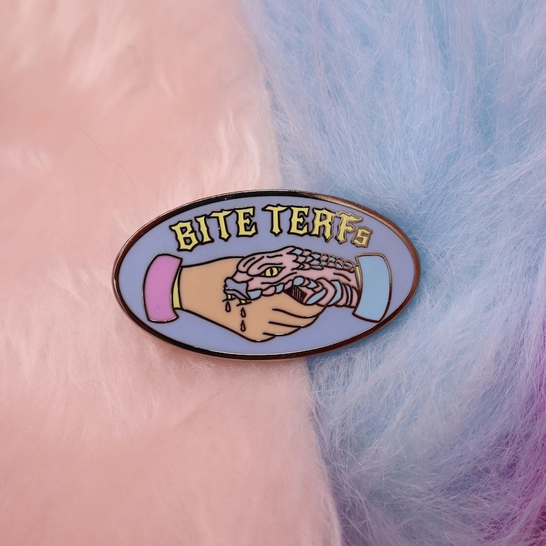 Puede incluir: Un pin de esmalte azul y rosa con una serpiente mordiendo una mano. El pin dice "Bite Terfs" en letras amarillas.