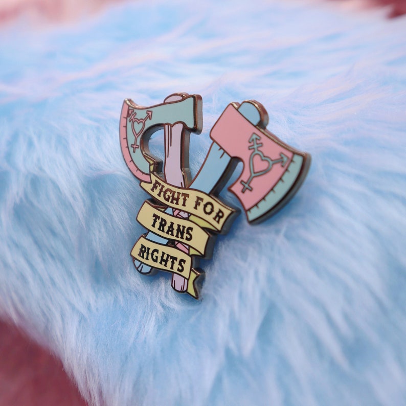 May include: A pink and blue enamel pin with two crossed axes and a banner that reads "Fight for Trans Rights".