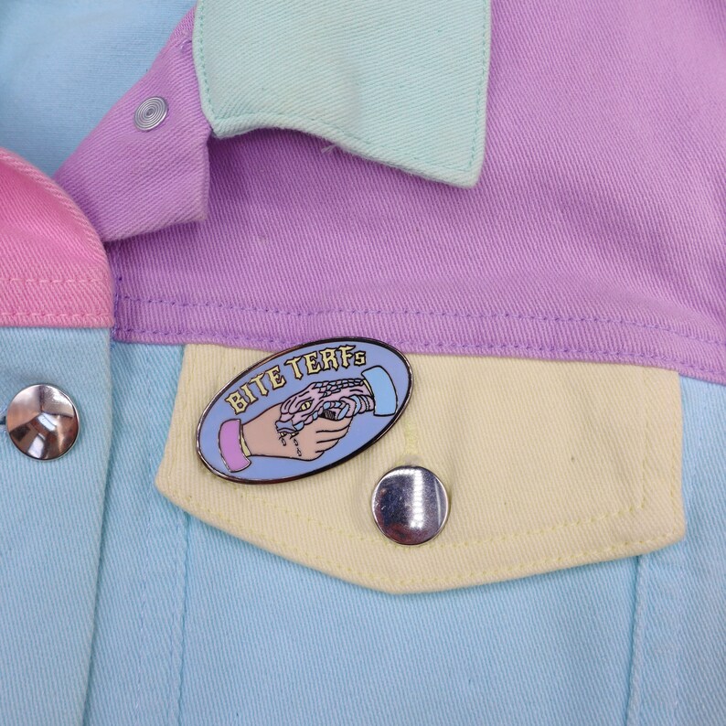 Puede incluir: Una chaqueta vaquera azul, amarilla, rosa y azul claro con un pin que dice "Bite Terfs" y muestra una mano estrechando la mano de una serpiente.