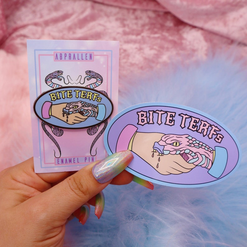 Puede incluir: Un pin de esmalte morado y azul con una serpiente mordiendo una mano. El pin dice "Bite Terfs" en letras blancas. El pin est&aacute; en una tarjeta blanca con las palabras "Abprallen Enamel Pin".