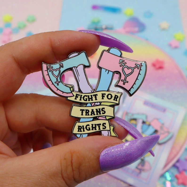 May include: A pink and blue enamel pin with two crossed axes, a heart with a transgender symbol, and the text "Fight for Trans Rights".