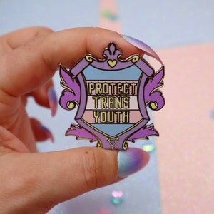 Pode incluir: Um broche de esmalte roxo, azul e rosa em forma de escudo com uma coroa no topo. O escudo tem as palavras "Protect Trans Youth" escritas nele. O broche tem um acabamento dourado e um símbolo transgênero na parte inferior.