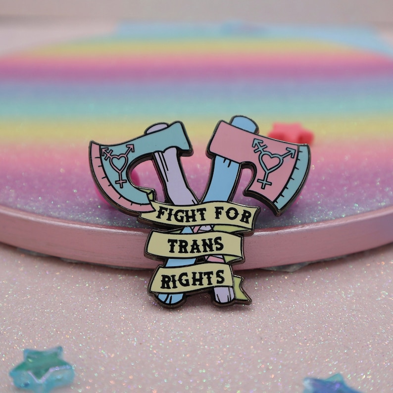 May include: A pink and blue enamel pin with two crossed axes, each with a transgender symbol. The axes are wrapped with a yellow ribbon that reads "FIGHT FOR TRANS RIGHTS".