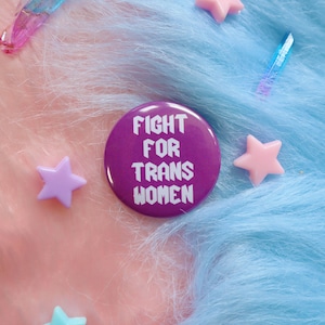Pode incluir: Botão roxo com o texto "FIGHT FOR TRANS WOMEN" em letras brancas. O botão está sobre um fundo de tecido felpudo rosa e azul.