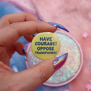 May include: A round, yellow button with the text "HAVE COURAGE! OPPOSE TRANSPHOBIA!" in blue. The button is held up against a pink and iridescent background.