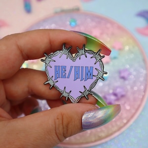 May include: A purple heart-shaped enamel pin with barbed wire around the edge. The words "HE/HIM" are printed in blue inside the heart.
