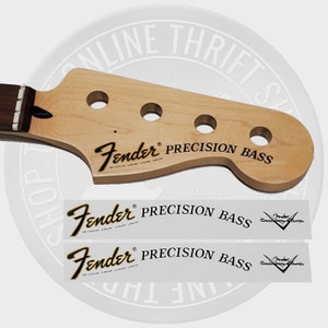 May include: A Fender Precision Bass headstock made of light wood, featuring three tuning holes and the Fender logo with "PRECISION BASS" in black. Two matching decals are below. The headstock is part of a musical instrument.