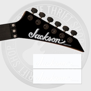 May include: Black guitar headstock with the brand name "Jackson" in white script. The headstock has six black tuning pegs and the text "MADE IN U.S.A." is visible. Two white stickers with the "Jackson" logo are below the headstock.