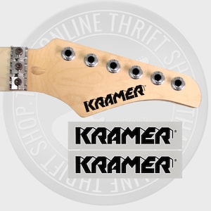 May include: Two black and white "Kramer" guitar headstock decals on a white background. The decals are rectangular and have a bold, stylized font.