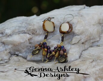 May include: A pair of dangle earrings with a white and brown square ceramic charm, gold beads, and brown tassels. The earrings are on a white rock.
