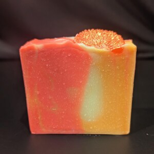 May include: A rectangular bar of handmade soap with a vibrant, swirled design in shades of orange, red, and white. A decorative orange slice is placed on top. The soap has a smooth, textured appearance.