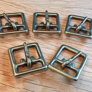 May include: Five square brass buckles with a rectangular frame and a central bar. The buckles are arranged on a light brown wooden surface. The metal has a shiny, aged appearance.