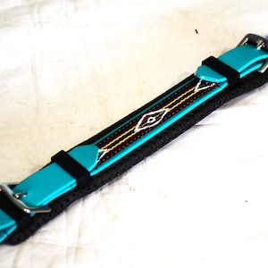 May include: A turquoise and black horse cinch with a decorative woven pattern. The cinch features turquoise straps with silver buckles and a black, textured center. The woven pattern includes white, orange, and black details.