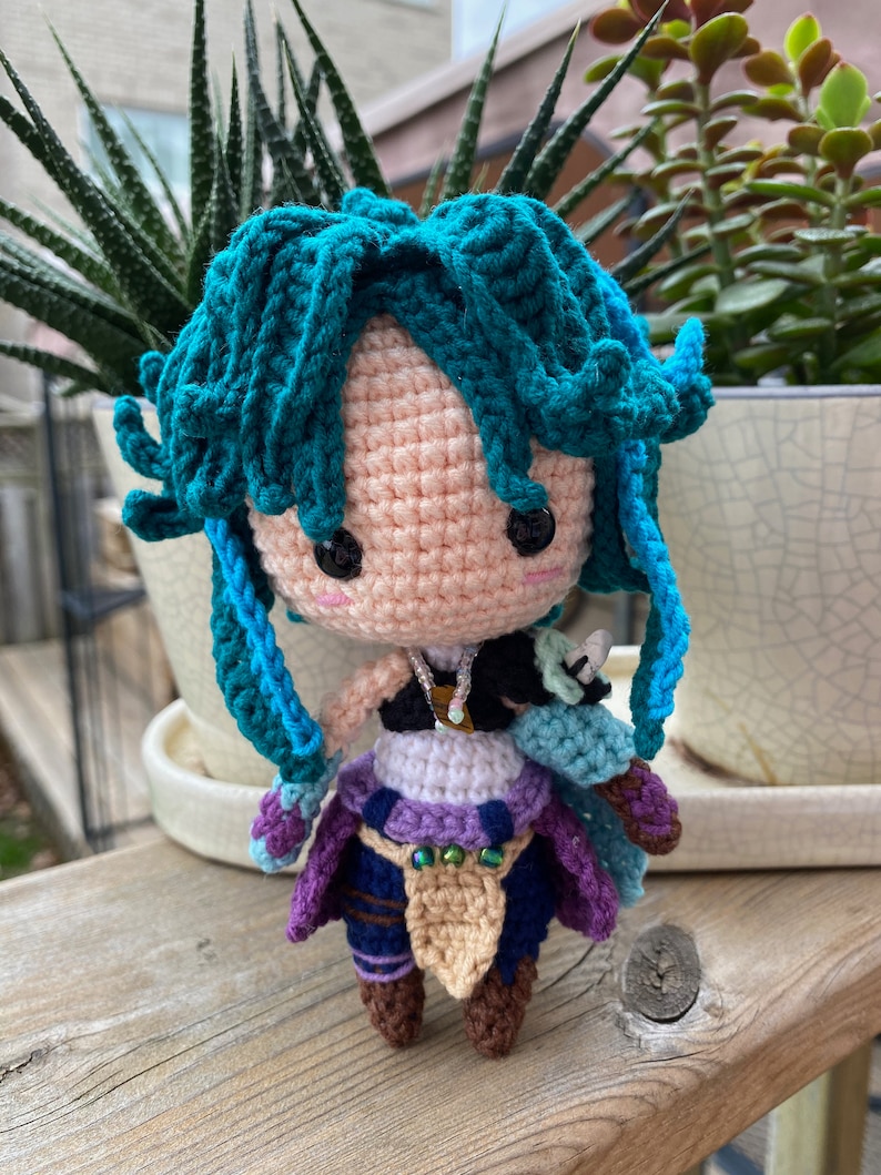 May include: Crocheted amigurumi doll with teal hair, a purple and blue outfit, and brown boots. The doll is standing on a wooden surface.