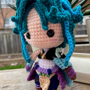 May include: Crocheted amigurumi doll with teal blue hair, a purple and blue outfit, and brown boots. The doll is holding a teal blue sword in its right hand.
