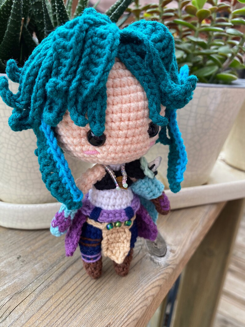 May include: Crocheted doll with teal hair, a purple and blue outfit, and brown boots. The doll is standing on a wooden surface.