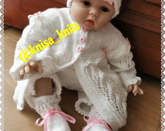 May include: A white knitted baby outfit with a matching hat and booties. The outfit features a delicate lace pattern and pink ribbon accents. The outfit is perfect for a newborn baby girl.