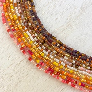 May include: A collection of colorful seed beads in shades of orange, yellow, brown, and pink. The beads are arranged in a fan shape, with the darkest colors at the top and the lightest colors at the bottom.