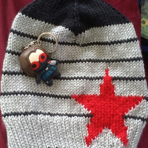 Puede incluir: Un gorro de punto de rayas grises y negras con una estrella roja y un pequeño llavero de personaje negro y rojo.