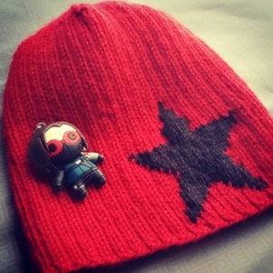 Puede incluir: Gorro de punto rojo con un diseño de estrella negra y un pequeño llavero de personaje negro y rojo.