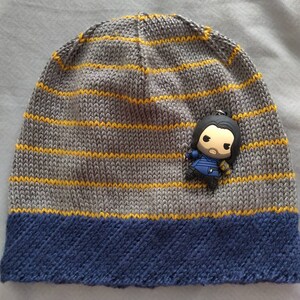 Puede incluir: Un gorro de punto de rayas grises y amarillas con un borde azul. Una pequeña figura de personaje está unida al gorro.