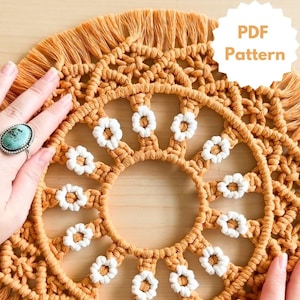 May include: A macrame wall hanging with a circular design featuring a fringe border and white daisy-like accents. The macrame is made with a mustard yellow cord.