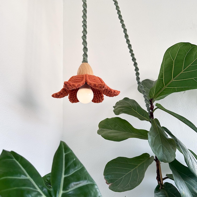 May include: A crocheted flower-shaped lampshade with a wooden base hangs from a green cord. The lampshade is a warm orange color and the light bulb is on.