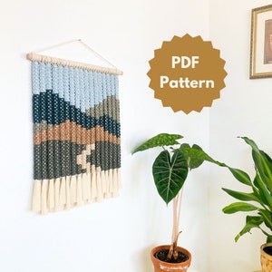 Op de afbeelding: Een wandkleed met een geweven patroon van blauwe, groene, bruine en witte garens. Het kleed heeft een franje van wit garen aan de onderkant. De afbeelding bevat ook een plant in een terracotta pot en een bord met de tekst "PDF Pattern".